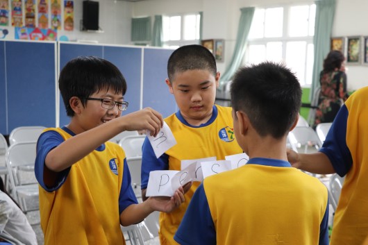教室內，穿著黃藍色運動服的小學生們聚集一起，其中一名學生正向其他同學展示並分發白色紙張或證書。背景可見教室布置、窗戶和其他學生，場景溫馨友善，呈現校園活動或頒獎典禮的氛圍。