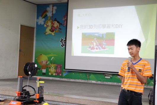 一位穿著橙色條紋衣服的男性講者在教室進行3D列印技術演講。講台上放置3D列印機，背景投影幕顯示中文標題「用我的3D列印學會和DIY」，牆上有卡通角色裝飾。