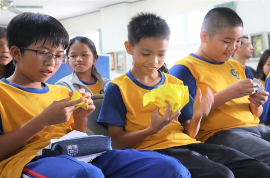 一群穿著黃藍色運動服的小學生坐在室內，專注地用手指觸摸和操作手中的物品。背景可見其他學生和室內環境，場景似乎是課堂或校內活動。