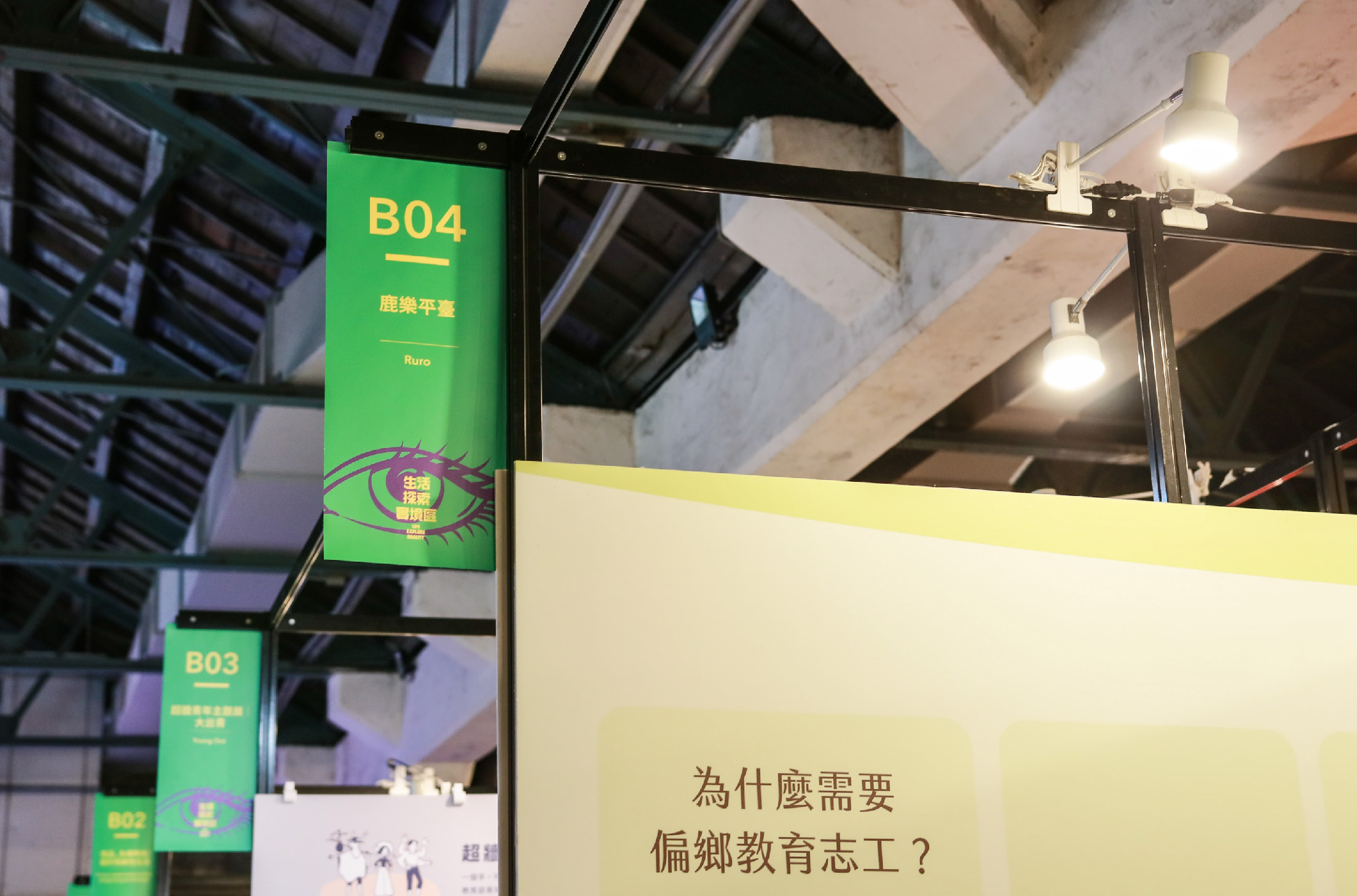 室內展覽場景，綠色標示牌顯示攤位號B04、B03、B02，標題為「鹿樂平臺」。右側黃色展板提問「為什麼需要偏鄉教育志工？」。天花板裝有照明設備，展現教育相關主題展示。