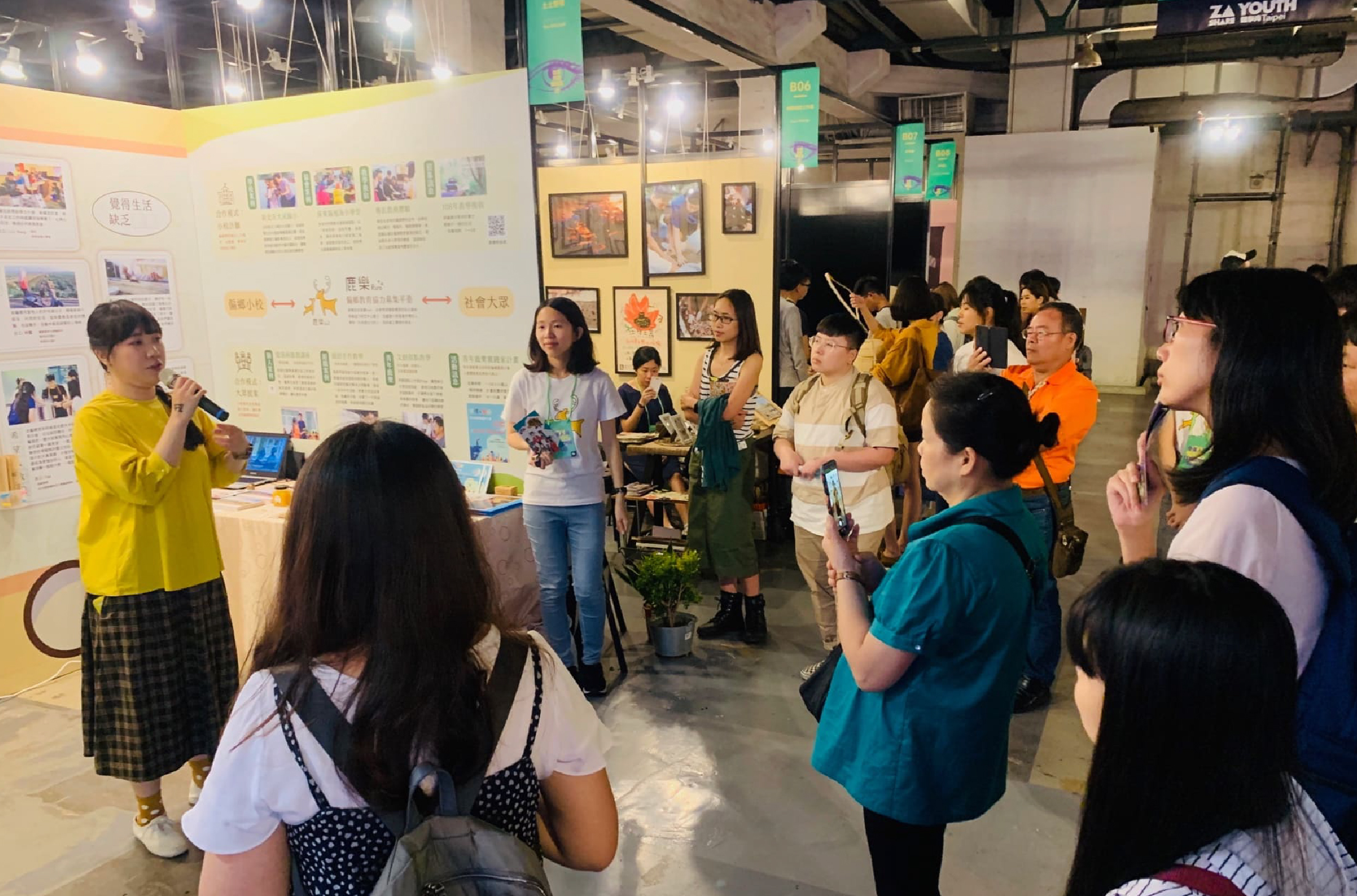 一位穿著黃色上衣的女性站在展示板前，向聚集的眾多參觀者介紹專案內容。現場展示多幅照片和文字說明，參與者認真聆聽簡介。這是一場在室內展廳舉辦的社區或青年活動展示會。