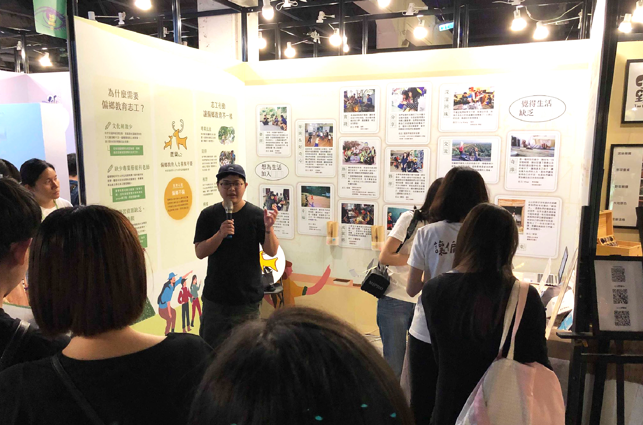 室內展覽現場，一位戴眼鏡的男性講者手持麥克風進行簡報，身後白色展板展示多張照片和中文文字說明。聽眾坐在前方聆聽，展板內容涉及社區生活與參與主題。