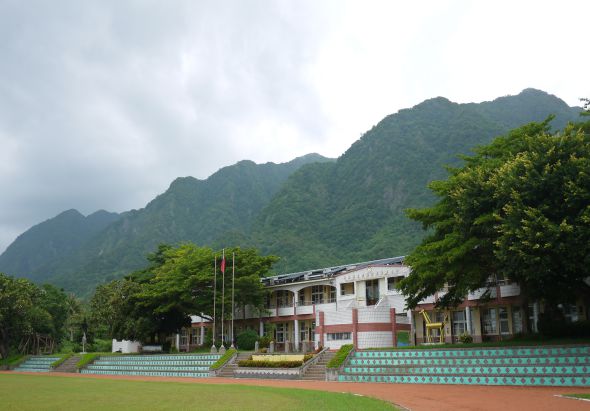 綠色山峰背景下，一棟紅白相間的多層建築物坐落於開闊的運動場旁。場地前方鋪設紅色跑道與綠色看台，周圍被茂密的樹木環繞，呈現學校或運動中心的建築景觀。