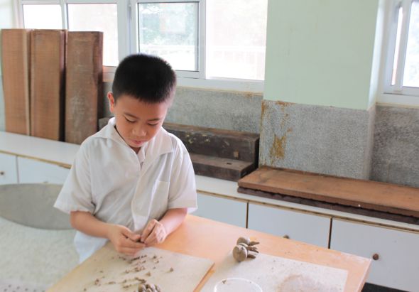 一名穿著白色短袖制服的男孩在教室內的工作台前，專注地進行手工藝製作。工作台上放著各種材料和工具。窗戶透進自然光，背景可見儲物櫃和建築材料。