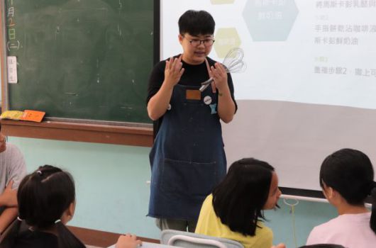 教室內，穿著黑色背心的年輕男性講師戴著眼鏡，在投影幕前向坐著的學生們進行教學。黑板和投影幕上有中文文字內容。學生認真聽講。