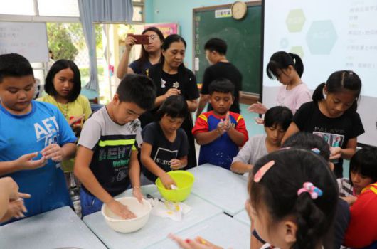 教室內一群小學生圍著白色工作台進行烹飪活動。中央學生正在攪拌碗中的食材，其他學生觀看或等待輪流。黑板、時鐘和教室布置清晰可見，展現出互動式的食品教育課程。
