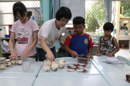 一位成人與四名兒童在教室內進行烹飪活動。他們圍在白色工作台前，正在準備食材和混合材料。桌上擺放著各種食材和烹飪用具，背景可見窗戶和教室布置。這是一堂實踐性的烹飪課程或工作坊。