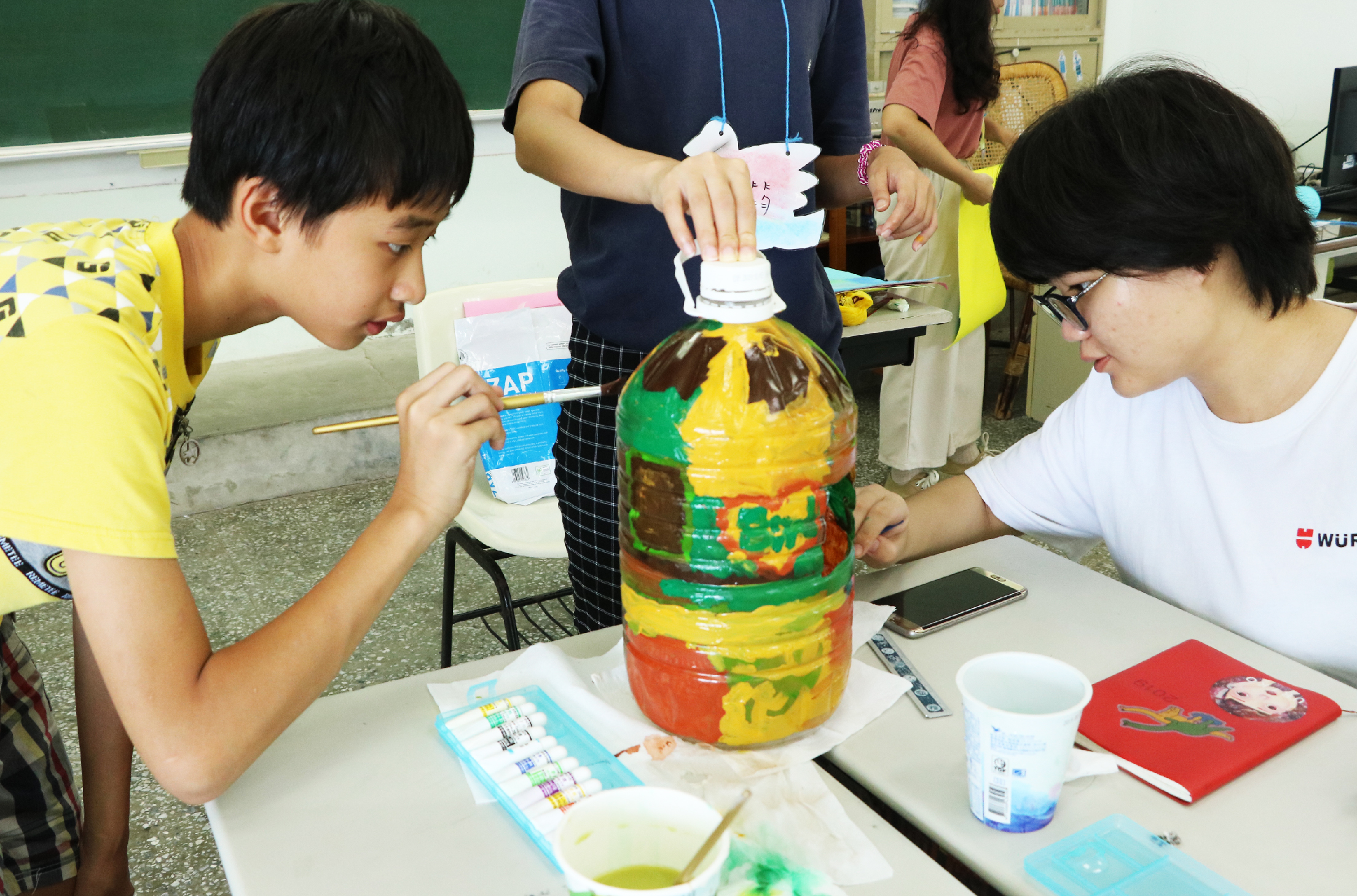 教室內，學生們正進行美術活動，用彩色顏料在大塑膠瓶上繪製圖案。左側穿黃色上衣的男生正認真作畫，右側戴眼鏡的女生也在繪製。中央站立的學生舉著完成的彩繪瓶作品，瓶身色彩繽紛。桌上擺放顏料盤、水杯與毛筆等繪畫工具。