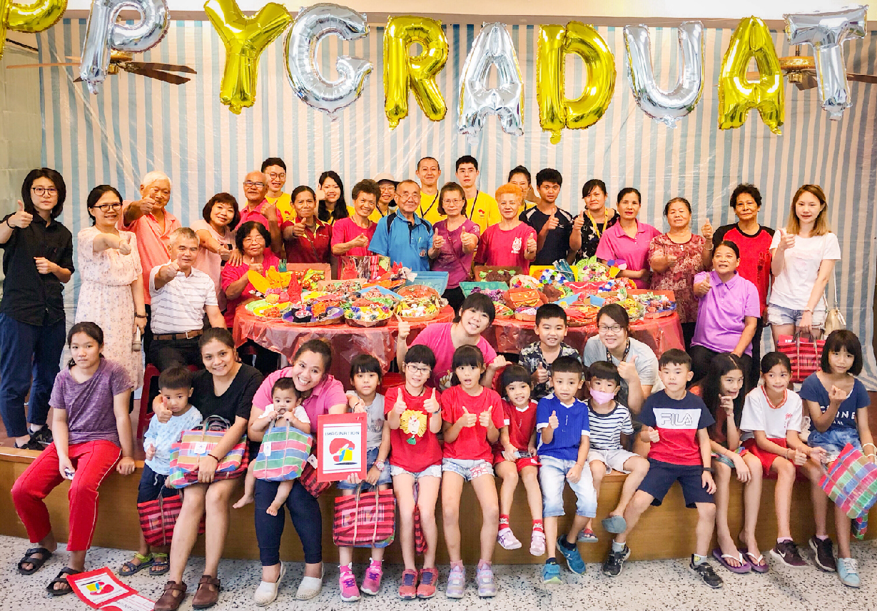 大群成人與兒童在室內聚集慶祝，背景懸掛黃銀色氣球拼成「YCRPDUA」字樣，桌上擺滿彩色食物與飲料，眾人做出歡樂手勢，呈現溫馨的集體活動氛圍。