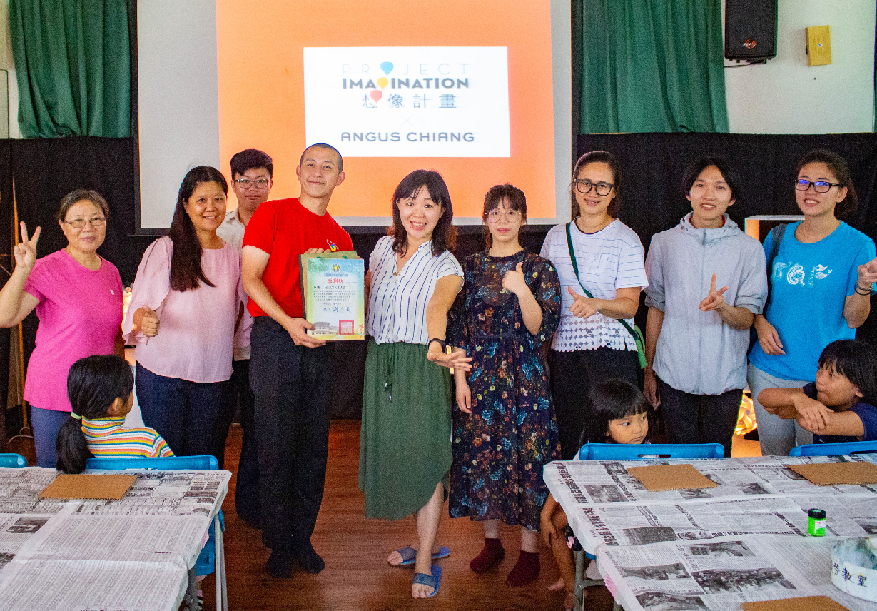 一群人在室內活動現場合照，背景展示「Imagination 想像計畫」及「Angus Chiang」標誌。男子穿紅衣持獎狀，女子穿著多彩，兒童坐在前方。桌上放置報紙，綠色窗簾和黑色舞台背景襯托溫馨氛圍。