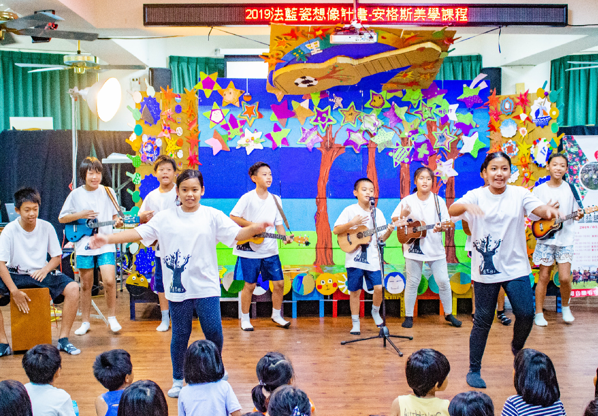 一群小學生在舞台上表演音樂，手持吉他和烏克麗麗。背景是色彩繽紛的星星和樹木裝飾，舞台上方掛著2019法藍瓷慈善音樂會橫幅。觀眾坐在舞台下方觀賞表演。