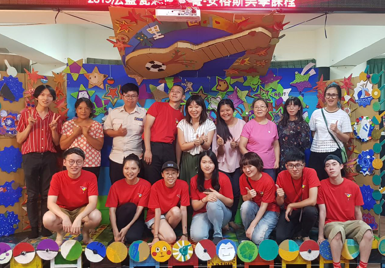 一群學生在校園活動現場合照，背景裝飾著五彩繽紛的紙質星星、吉他造型和中文字樣。前排學生穿紅色制服跪坐，後排學生站立穿著各色服裝。現場布置充滿節慶氣氛，展現校園文化活動的歡樂氛圍。
