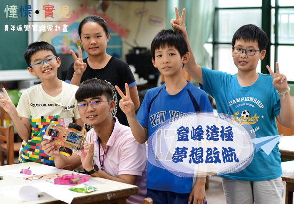 六位小學生在教室內舉著寫有「啟發港孩 夢想飛航」的標語牌，展示自製的彩色積木和手工作品，開心地做出勝利手勢，展現創意學習成果的校園活動場景。