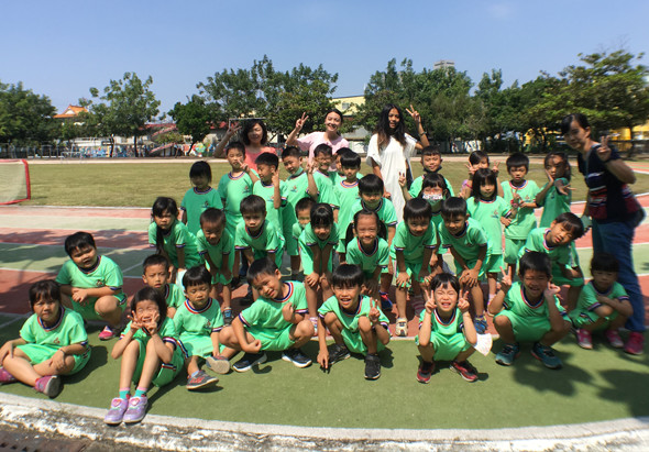 一群穿著綠色運動服的小學生在戶外運動場集合拍團體照。孩子們分為前後排坐姿和站姿，背後是教練和老師站立。場景為晴朗的校園操場，遠方可見樹木和校舍建築。