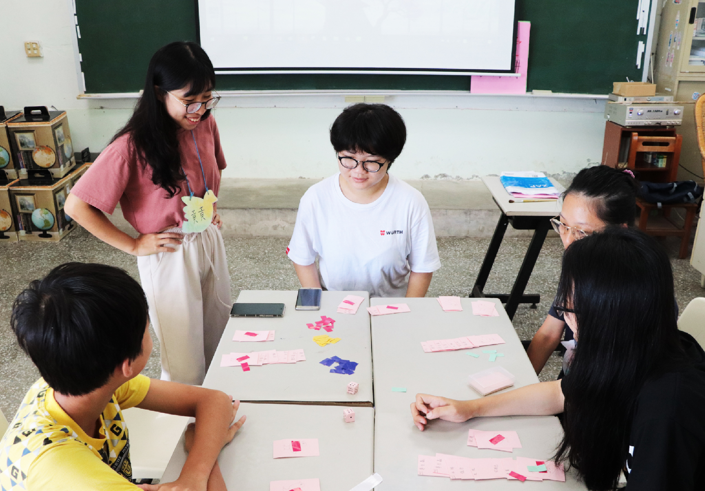 教室內，一位女性教師站在講桌旁，與四名學生圍成一圈進行互動教學。桌上散放著粉紅色和彩色紙張、便籤紙及學習教具。黑板和教室物品可見於背景，呈現活潑的課堂學習氛圍。
