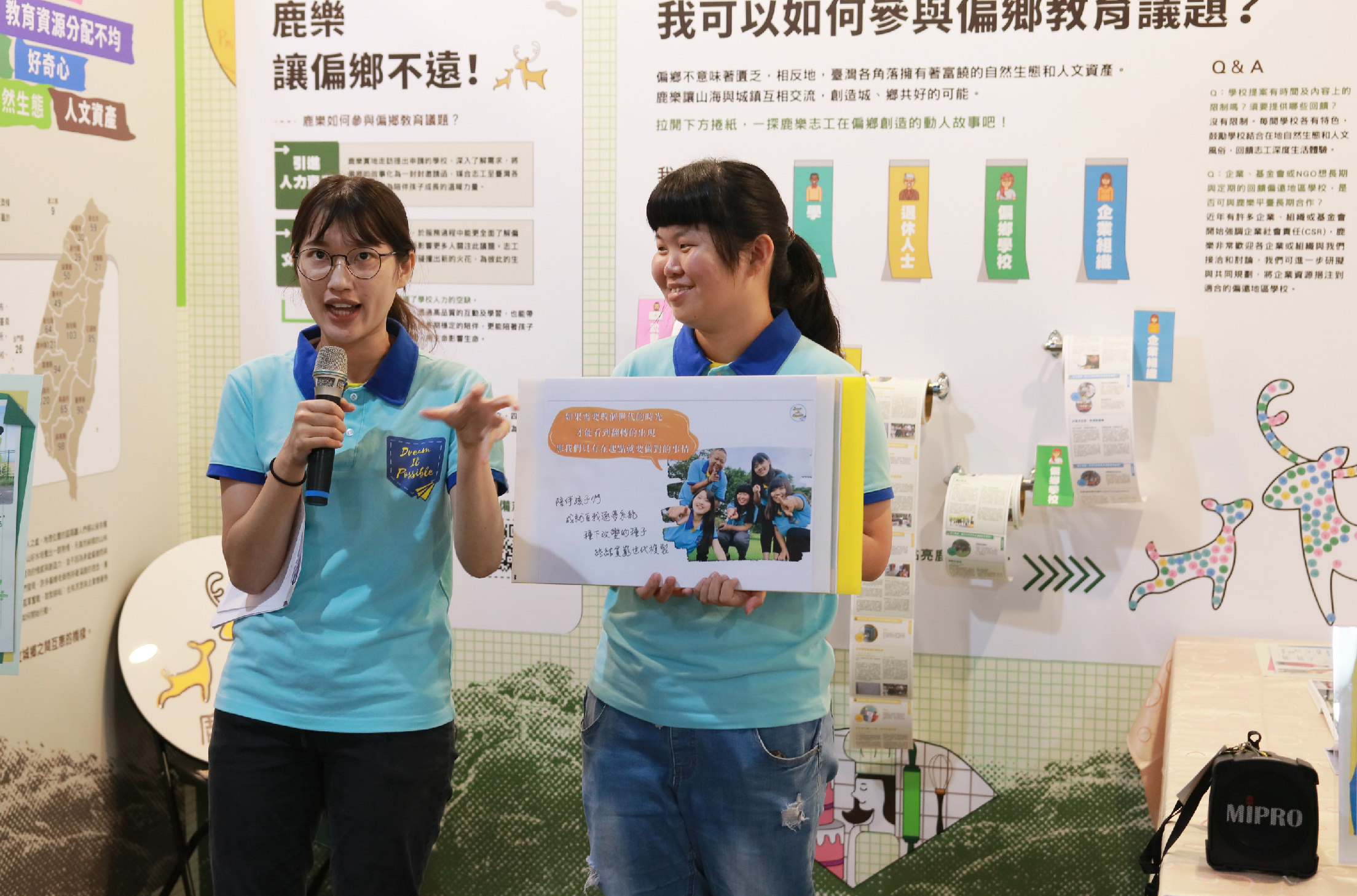 兩名穿著藍色polo衫的女學生在教室進行簡報，左邊學生手持麥克風說話，右邊學生展示海報。背景牆面貼滿與鄉村教育相關的宣傳海報和彩色裝飾，地上放有黑色音響設備。
