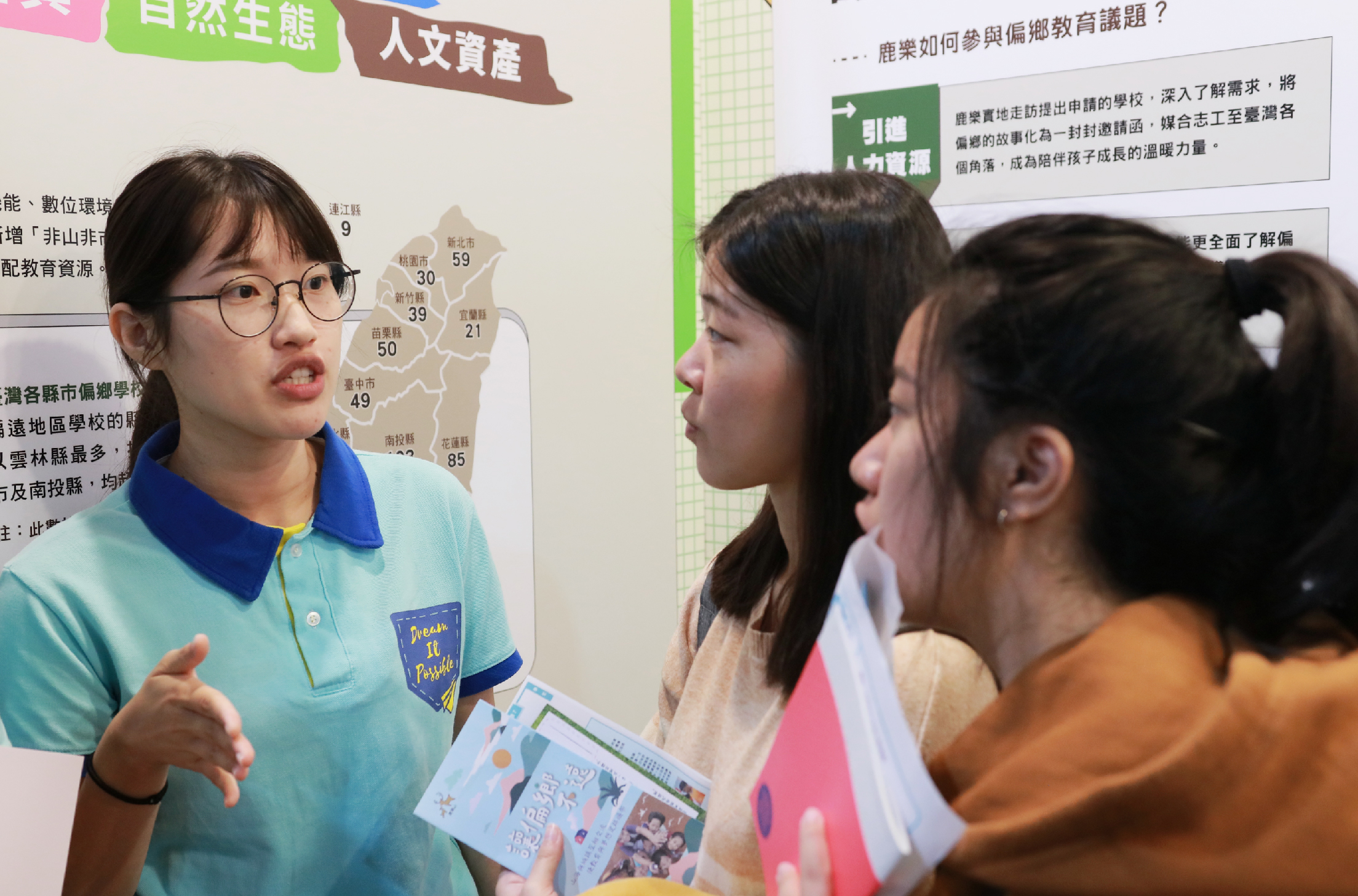 一名戴眼鏡穿著藍綠色polo衫的女性講者，在布告欄前向三名學生介紹教育議題。背景展示台灣地圖及相關統計資訊，講者手持教材進行說明。