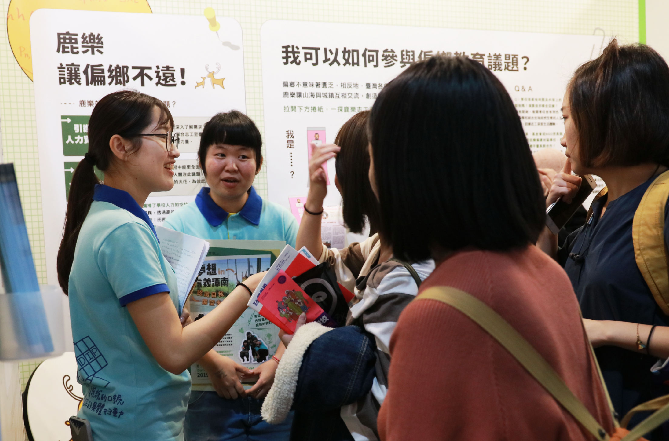 教育推廣活動現場，兩位穿著藍色polo衫的年輕講者正向參觀者介紹展示板。展板標題為「鹿樂讓偏鄉不遠」和「我可以如何參與作伙來議題」，講者手持宣傳資料與訪客互動交流，現場聚集多位感興趣的參觀者。