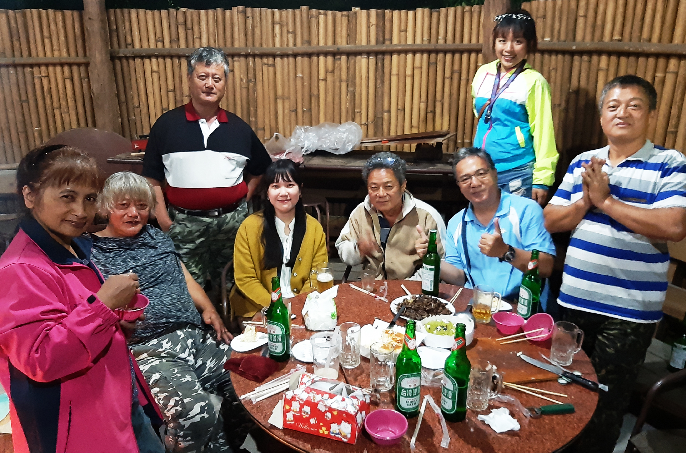 一群人在竹製建築物內聚餐，圍坐在木製餐桌周圍。桌上擺放啤酒瓶、食物和餐具。眾人穿著色彩繽紛的衣服，有人舉起啤酒瓶做出祝賀手勢，展現歡樂的聚會氛圍。背景是竹編牆壁。