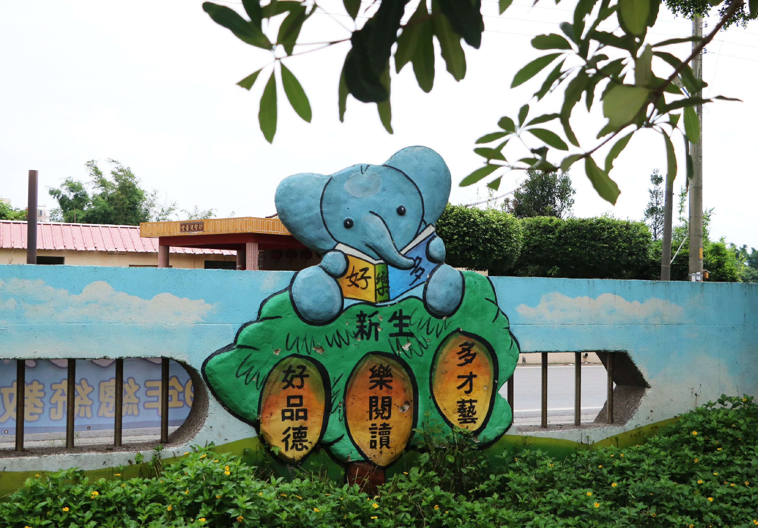 校園牆面彩繪，描繪一隻藍色大象坐在綠色樹葉上，象身穿著黃色衣物，樹葉下方三顆橙色果實上書有中文字句，宣傳品德教育和才藝培養的校園標語。