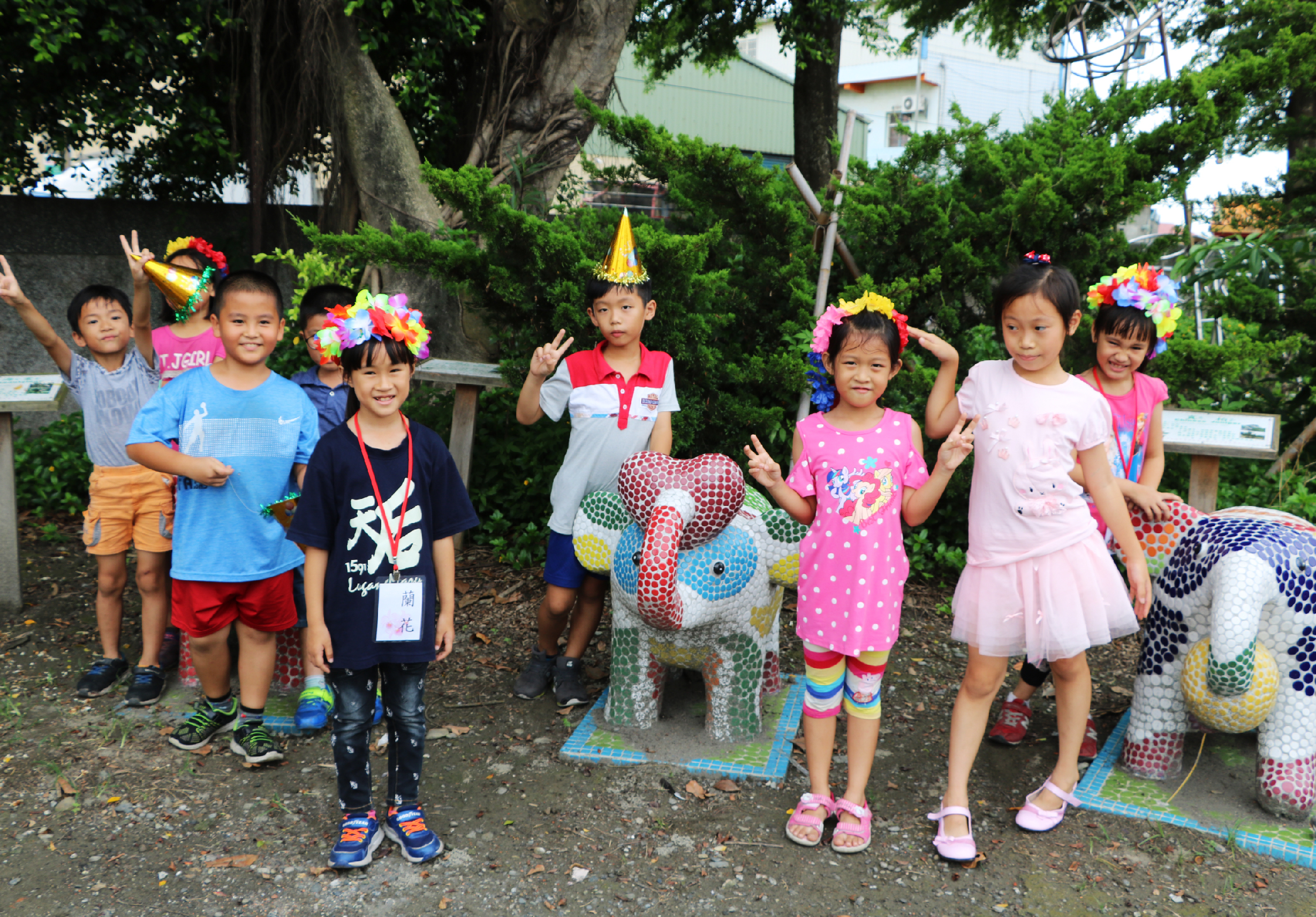 一群小學生在戶外草地上歡樂慶祝，戴著彩色花圈和派對帽，穿著五彩繽紛的衣服。他們圍在用彩色網格裝飾的大恐龍造型物件周圍，開心地擺姿勢拍照，背景是大樹和校園建築。