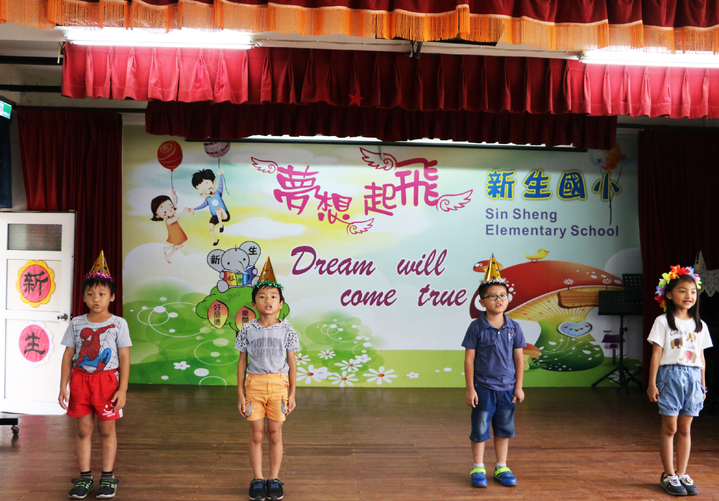 新生國小學生在舞台上進行表演。四名小學生穿著彩色服裝，戴著花冠站成一排。舞台背景為五彩繽紛的海報，寫著「夢想起飛」、「Dream will come true」等英文和中文標語，裝飾著紅色和金色的舞台帷幕。