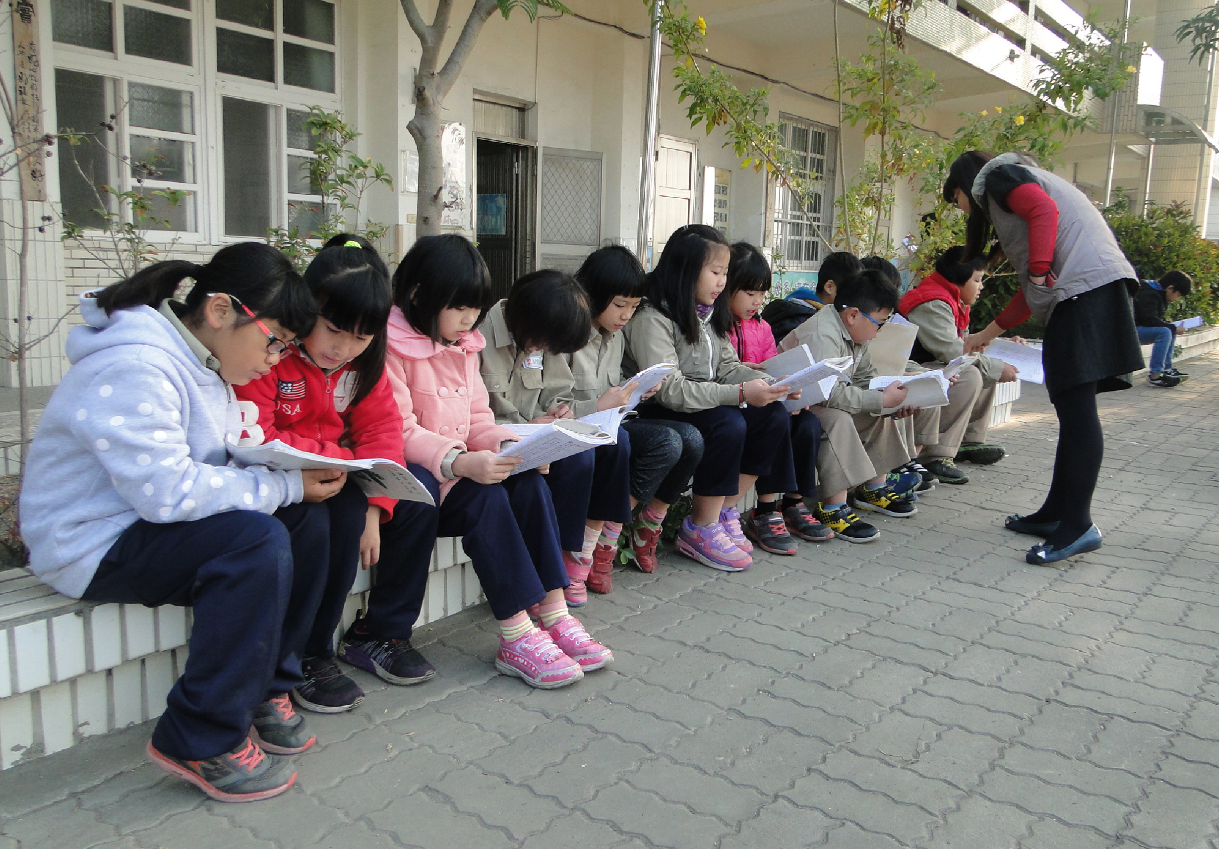 一群約十多位小學生在學校走廊邊排成一列坐著，低頭認真閱讀手中的紙張。成人站在旁邊監督。孩子們穿著冬季外套，背景是校舍建築和綠色植物。