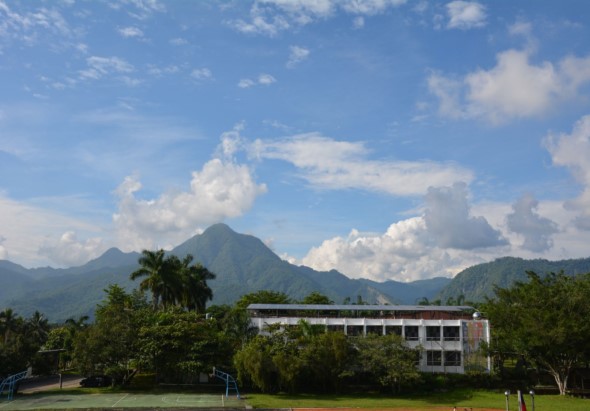 白色現代建築物坐落在綠色草坪上，背景是聳立的綠色山峰，藍天白雲映襯著遠方山脈。前景有棕櫚樹和各種綠色植被，呈現典型的熱帶山區風景。