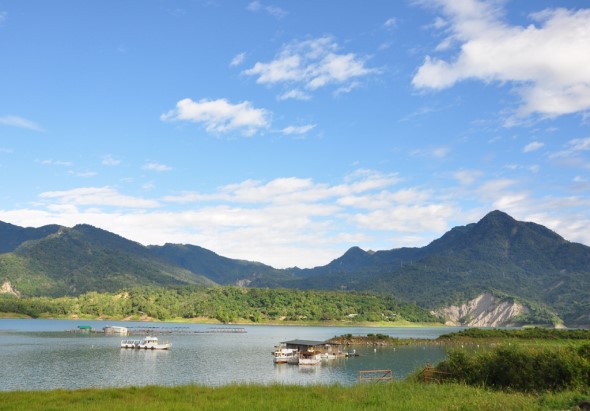 寧靜的湖泊風景，清澈碧綠的水面上停泊著幾艘小木船。背景是連綿的青山，綠色植被覆蓋山坡，右側岸邊有陡峭的土質斜坡。前景是翠綠的草地，藍天白雲映照水面，呈現自然和諧的生態景觀。
