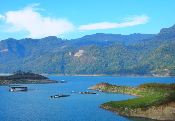 青山環繞的寬闊藍色湖泊，湖面上有數艘小船停泊，前景為綠色半島，背景為層層疊疊的翠綠山脈，晴朗藍天飄著白雲，構成寧靜秀麗的山水風景。