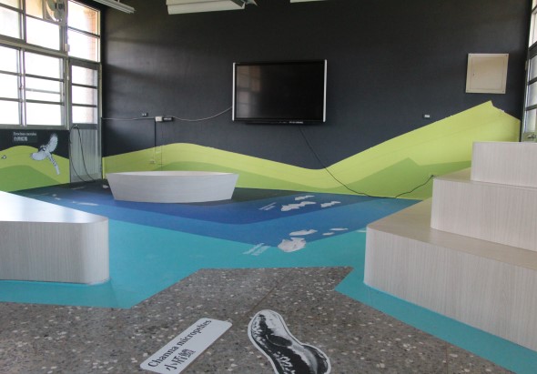 室內展示空間，地面以藍色代表水域，灰色代表河床，綠色波浪形牆面代表陸地，展示生態環境主題。牆上掛有電視螢幕，白色圓形和方形裝置作為展示台。地上放置鞋類物品和標籤。左側窗戶採光充足。