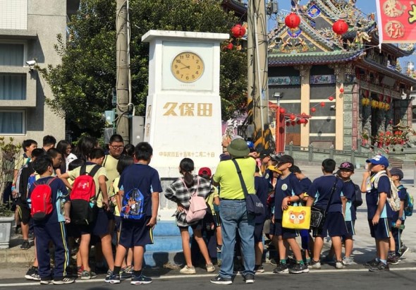 一群穿著校服的學童在街道上集合，聚集在寫有「久保田」字樣的白色時鐘標誌前。背景可見中式廟宇建築、紅燈籠和裝飾。一位穿著黃色安全背心的成人在引導學童，可能是學校外出參訪或戶外教學活動。