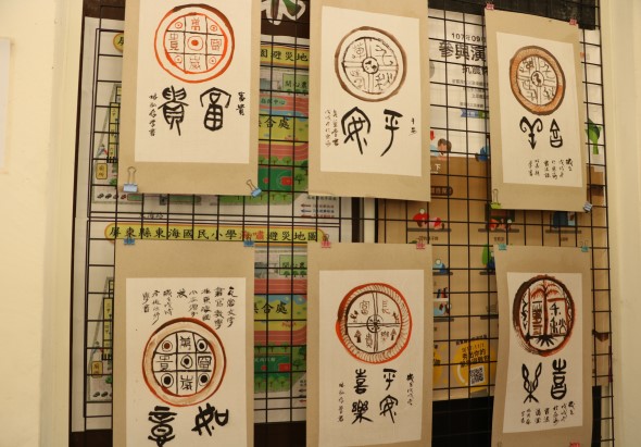 展示牆上貼著六張宣紙，每張均繪有紅色圓形印章圖案和書法文字。印章內含八卦符號、文字或圖騰，搭配毛筆書寫的篆書和隸書。背景為方格網面。這些作品呈現傳統中文書法和印章設計藝術。