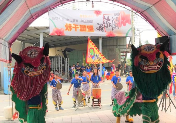 廟會活動現場，兩隻紅綠色的大型獅頭擺設在拱形帳篷入口兩側。舞台上穿著藍色上衣的表演者正進行傳統舞獅表演，觀眾圍觀欣賞。上方掛著紅色活動橫幅和裝飾旗幟。
