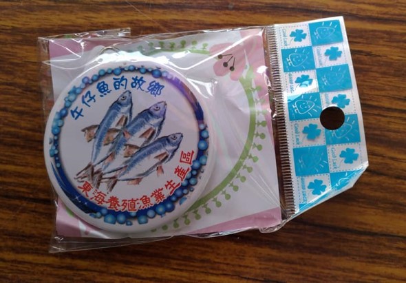 塑膠包裝內放置圓形瓷盤，盤面繪有三尾魚的藍白青花瓷圖案，外圈飾以藍色珠紋邊框。盤底有紅色印章。包裝右側附藍白相間的棋盤花紋紙質標籤。木製背景上展示。