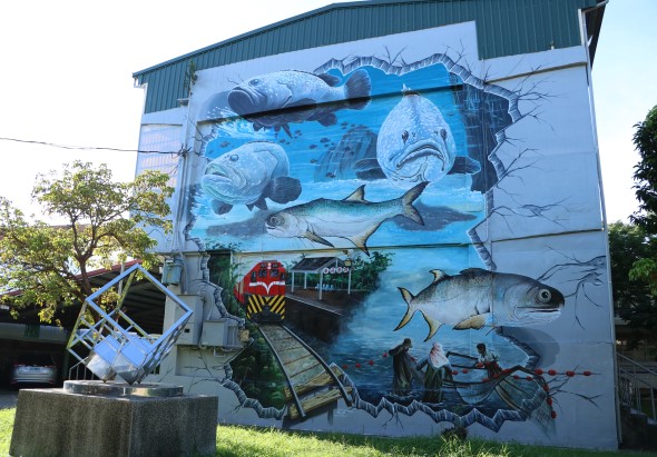 工業建築外牆上的大型海洋生物壁畫，繪有鯊魚、魟魚、箭魚等海洋生物，色調以藍色為主。建築物旁停放著紅色列車模型，地面有綠色草坪和樹木。