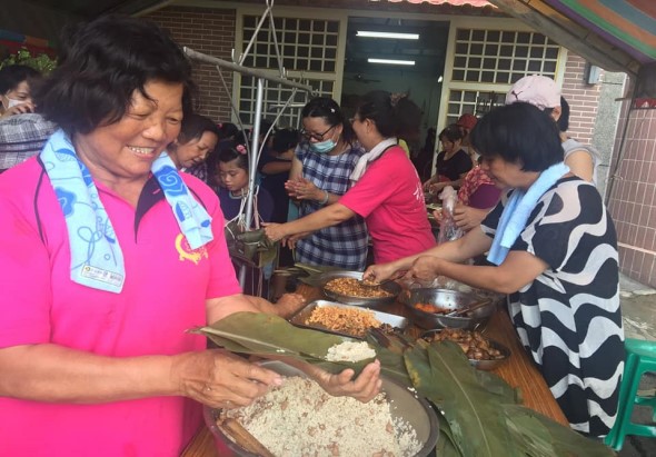 一場社區活動現場，多位佩戴藍色緞帶的志工在室內為民眾盛裝食物。前景中穿著粉紅色上衣的志工正用香蕉葉盛裝米飯，後方志工則在分發其他菜餚。現場擁擠，參與者眾多，氣氛熱鬧溫馨。