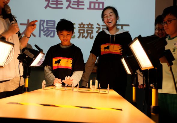 一群年輕人在舞台上進行科學實驗展示。中央的女性和男孩在實驗台前操作，周圍的人舉著照明燈具進行照亮。背景投影幕顯示中文字幕。參與者穿著印有西班牙國旗圖案的黑色T恤，現場氛圍熱烈歡樂。