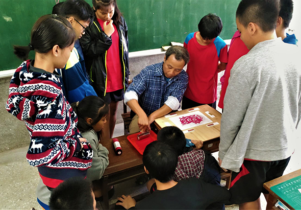 教室內，一位男性講師在講台前向圍成一圈的十多位學生演示剪紙或手工藝作品。綠色黑板背景下，學生們認真觀看講師手中的紅色紙製品，專注參與這場互動式的手工藝教學活動。