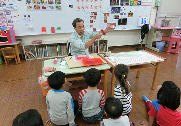 一位男性教師在教室裡向圍坐成半圓形的六位小學生進行教學。講師坐在桌前，手持教材指向黑板。教室牆面張貼著學生作品和教學資源。書架和粉紅色儲物櫃位於教室兩側。
