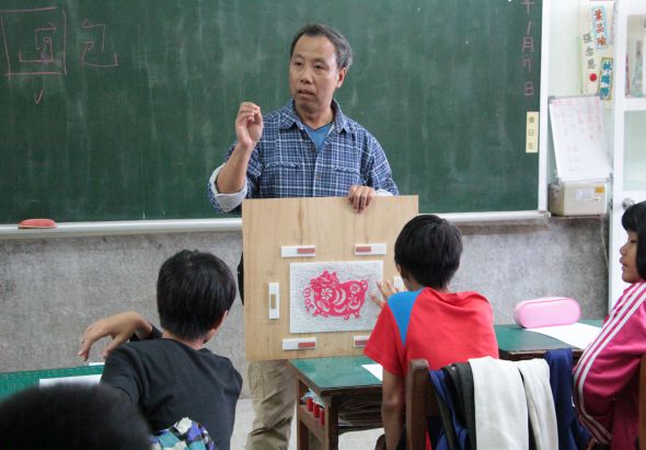 教室裡，一位男性教師站在綠色黑板前，手持教學用具向學生講解。他身前展示著一個木製教具盒，上面貼有紅色圖案。四名學生坐在課桌前認真聽講，教室牆上貼有各色學習資料。