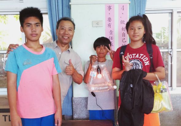 四名青少年在教室內合照，背景白板上寫著中文字。左起為穿著藍粉色T恤的少年、穿著淺色襯衫的成人、穿著灰色T恤的少年，以及穿著紅色T恤背著黑色背包的少女。人物面帶笑容，氛圍友善溫馨。