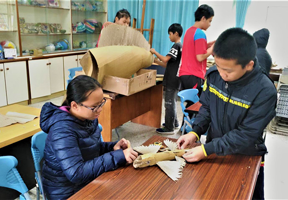 教室內一群學生正進行戶外教育活動。前景兩名學生在木桌上觀察並處理螃蟹標本，背景其他學生圍觀或操作教材。牆上有展示櫃和藍色窗簾，地板鋪有黃色墊子，營造互動式學習環境。