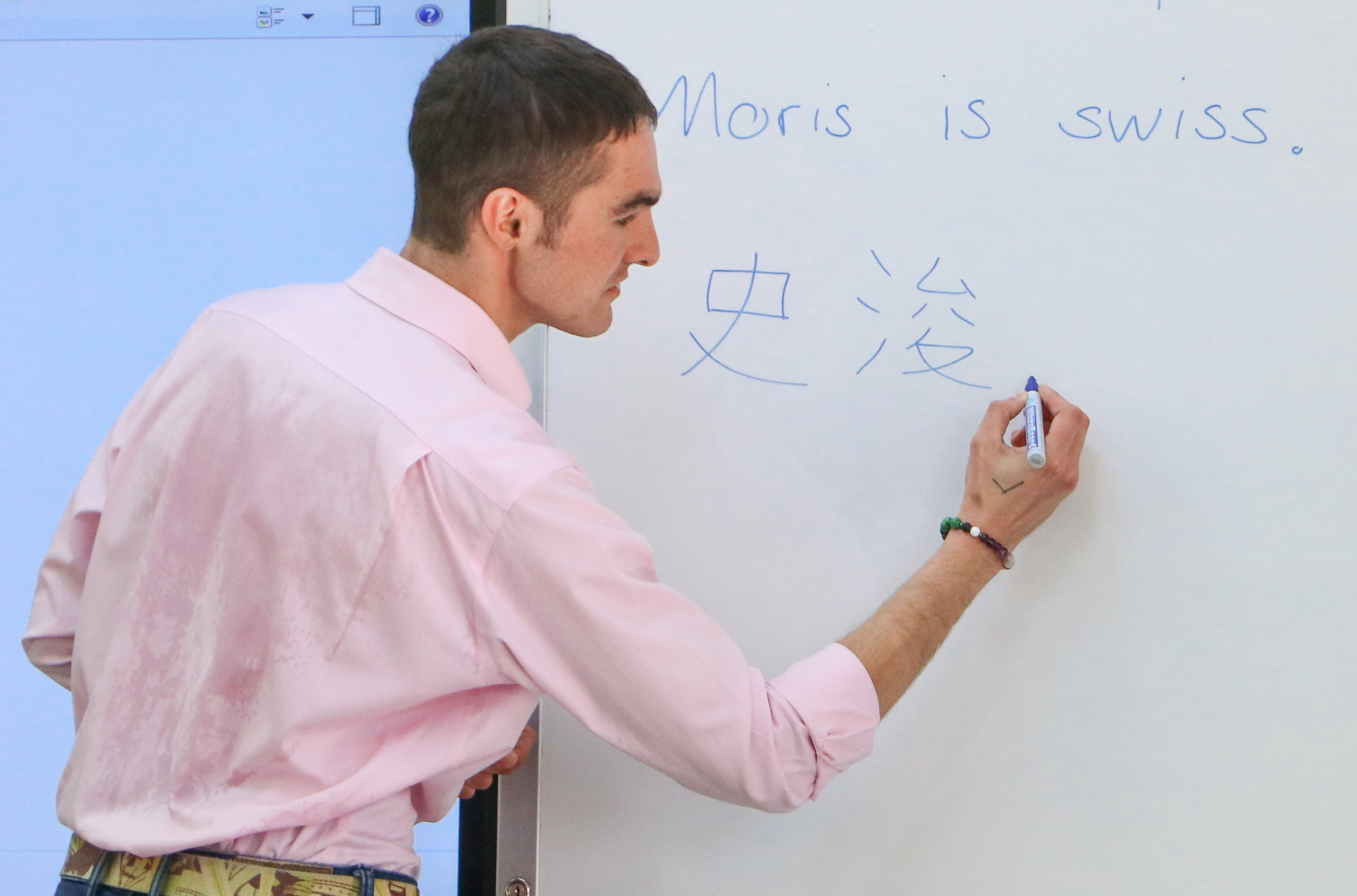 穿著粉紅色襯衫的男性教師正在白板上書寫，板上寫有「Moris is swiss」英文文字和中文漢字。他手持藍色筆，側身面向白板進行教學。