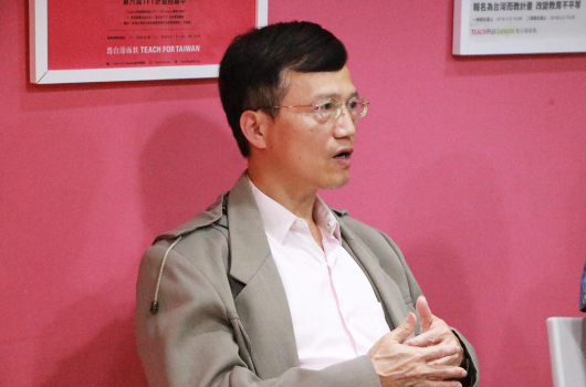 一位戴著眼鏡、穿著灰色西裝和白色襯衫的男性講者，在粉紅色背景的室內環境中進行演講或教學，身體呈側面，雙手做出說明手勢，背景牆上貼有紅色海報。