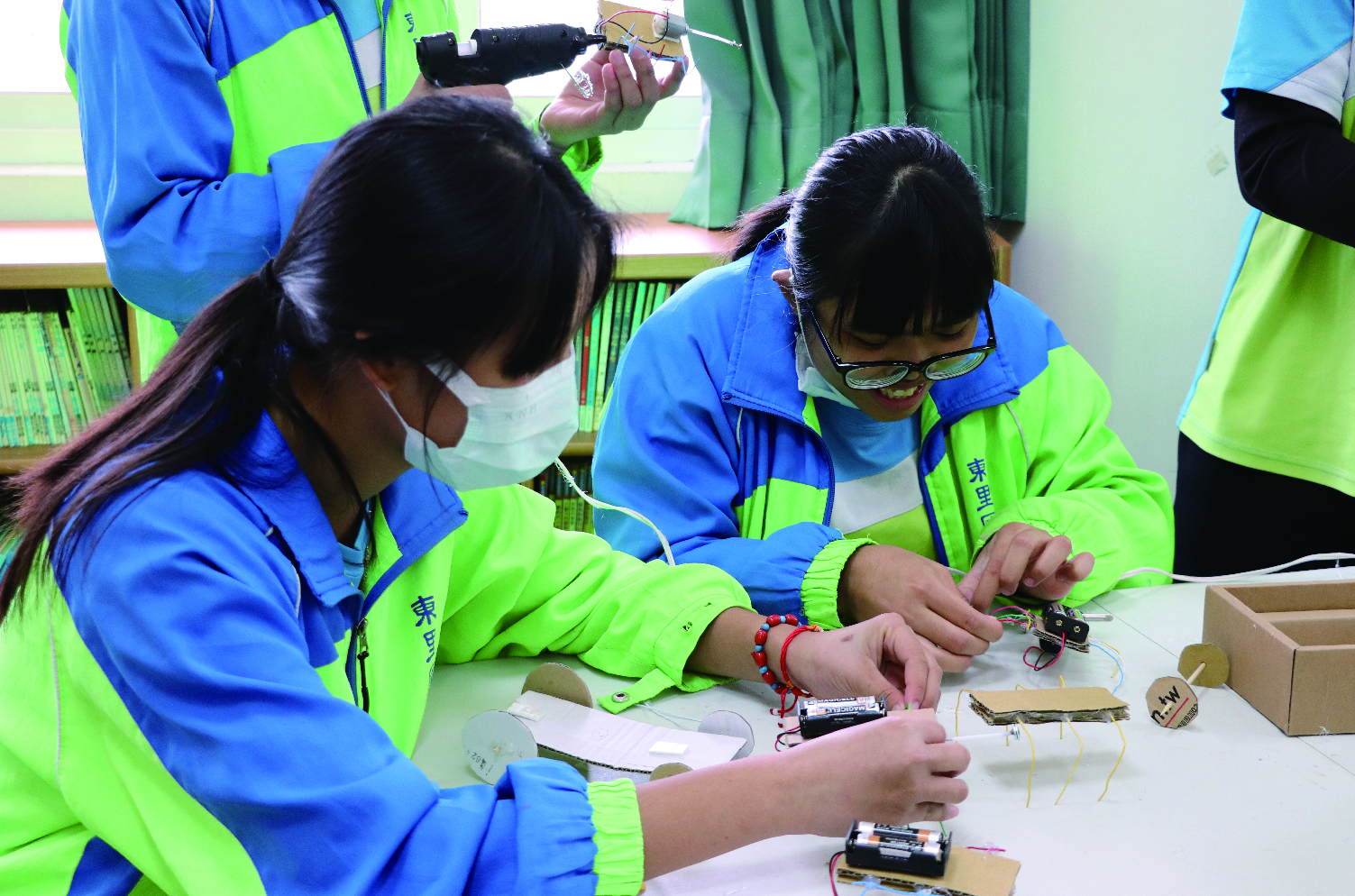 穿著藍綠色制服的學生在室內進行科學實驗課程，他們正在組裝電路和電子元件。學生們聚集在工作台前，操作電池、電線和傳感器等物品。背景可見書架和教學布置，展現動手科技學習的場景。