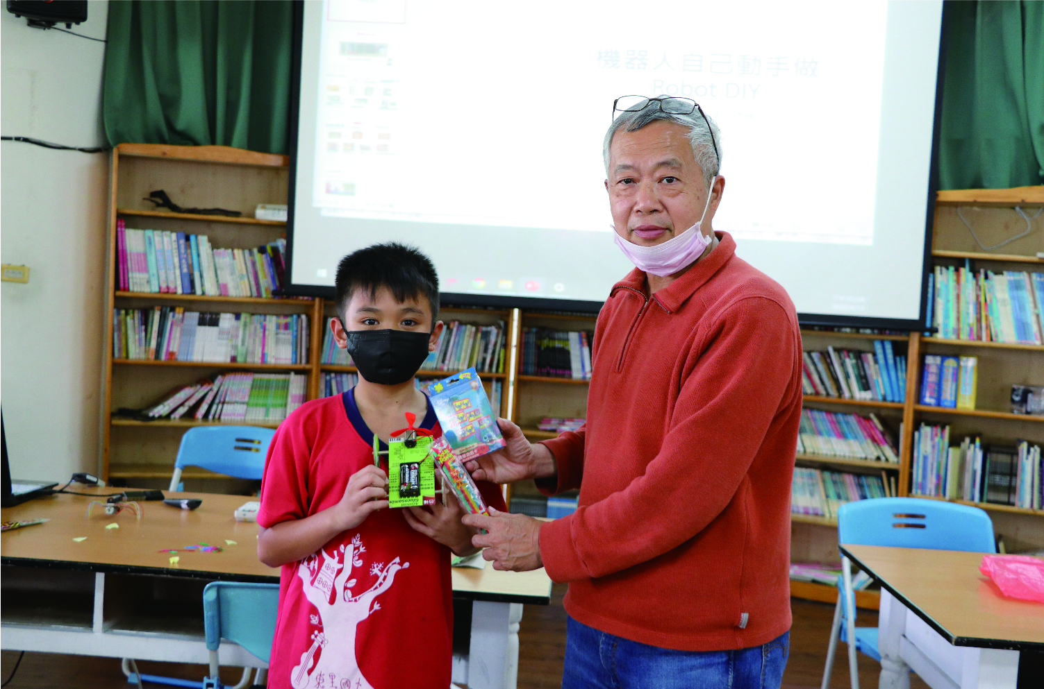 學校圖書館內，一位年長男性穿著紅色外套戴著紫色口罩，正向穿著紅色上衣戴著黑色口罩的小男孩頒發獎品。背景為木製書櫃、投影幕和綠色窗簾，桌上放有學習用具。