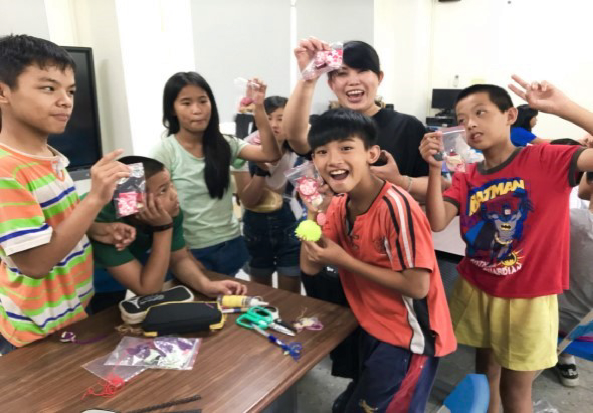 一群小學生在室內圍聚在木桌旁，歡樂地展示手中的粉紅色手工藝品和獎牌。孩子們穿著色彩繽紛的服裝，開心地笑著，桌上散放著製作材料和工具，呈現充滿活力的課堂活動或工作坊場景。