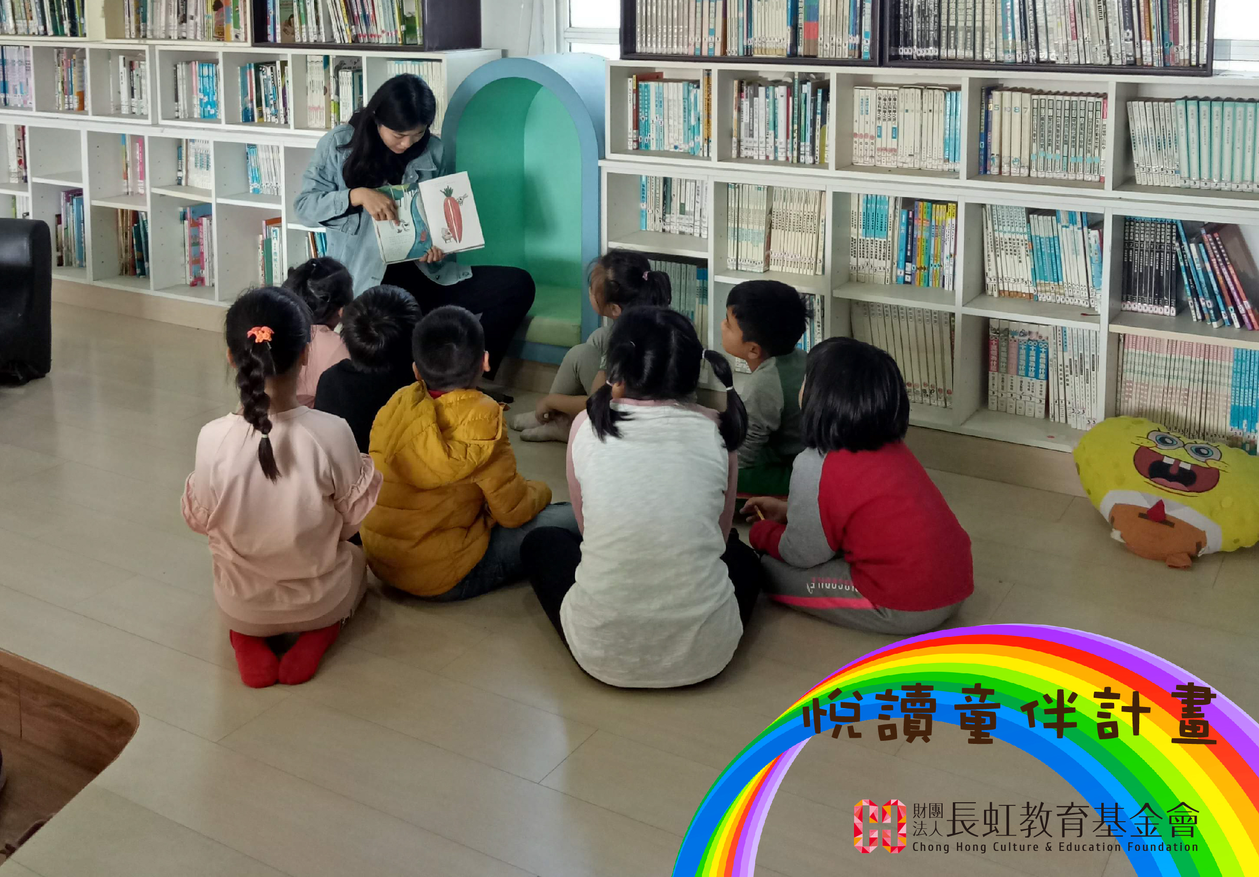 圖書館內，一位女性講師坐在綠色帳篷前，向圍坐成圈的六位幼童展示繪本。書架背景擺滿各式書籍。前景彩虹拱形標誌寫著「悅讀書伴計畫」，標示長中町教育基金會。