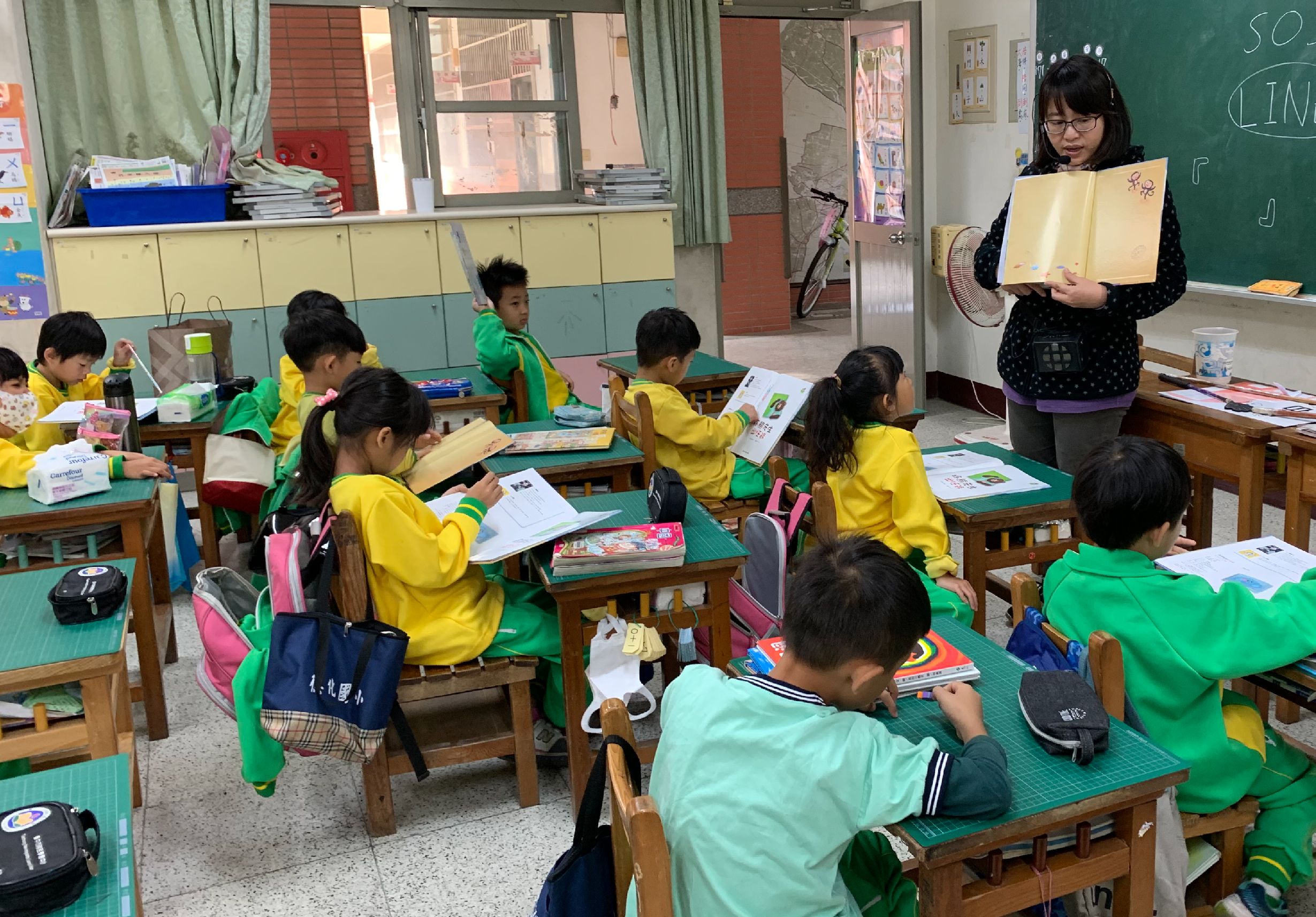 小學教室內，穿著黃綠色校服的學生坐在木製課桌前專心學習。女教師站在講台旁展示黃色教材，黑板、書櫃和窗戶可見。教室布置溫馨，學生認真聽講。